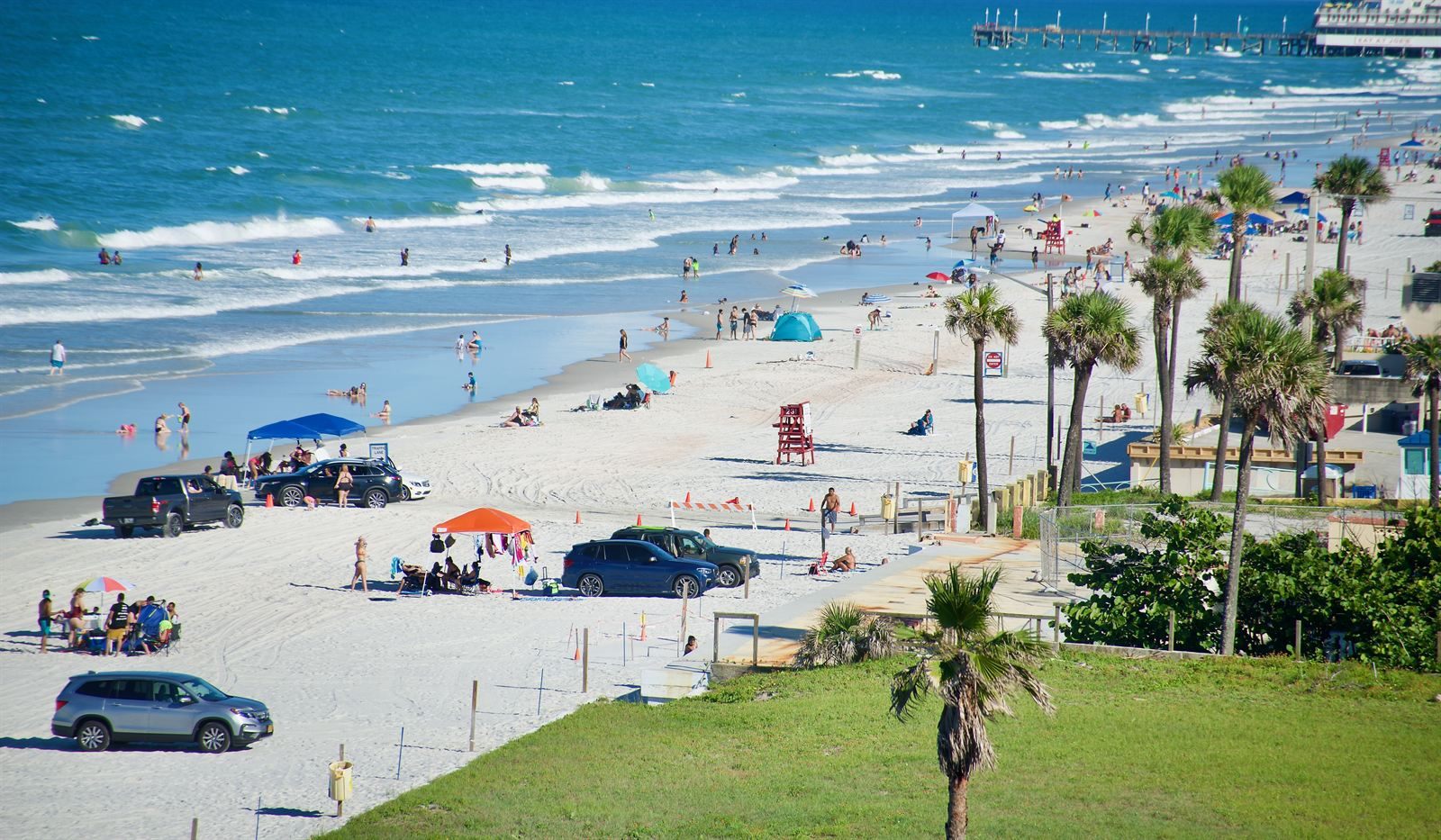 Cars and sun shelters parked on a wide beach of hard-packed sand, with palm trees and the surf beside them